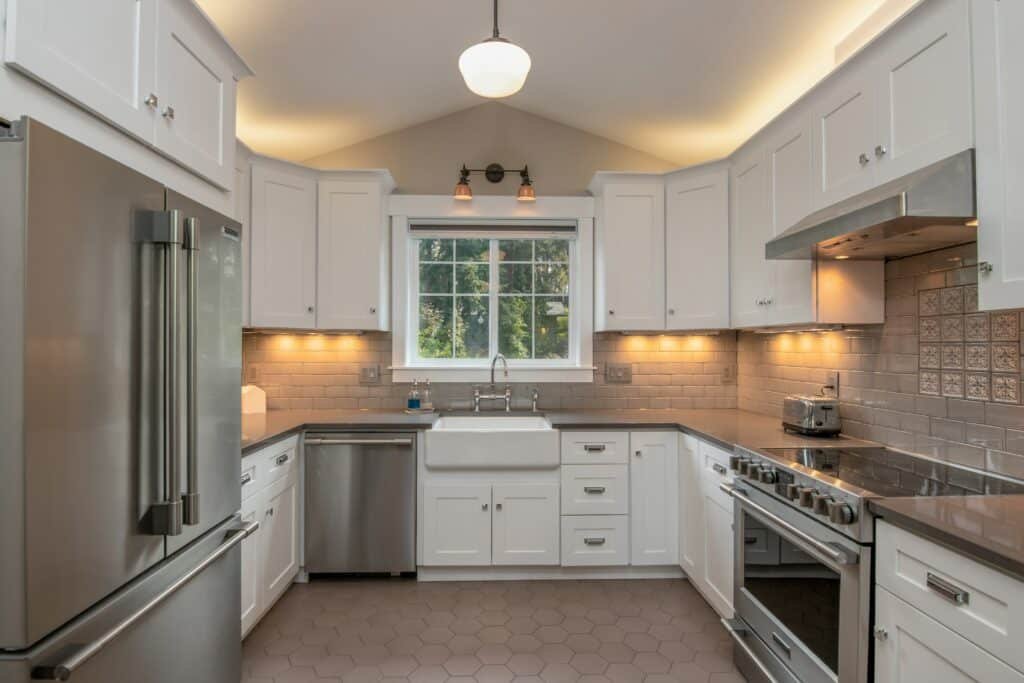 Traditional kitchen with white shaker cabinets, a farmhouse apron sink, stainless steel appliances, and a taupe subway tile backsplash. The space features warm under-cabinet lighting, vaulted ceilings with ambient cove lighting, and hexagon tile flooring for a blend of classic and modern design.