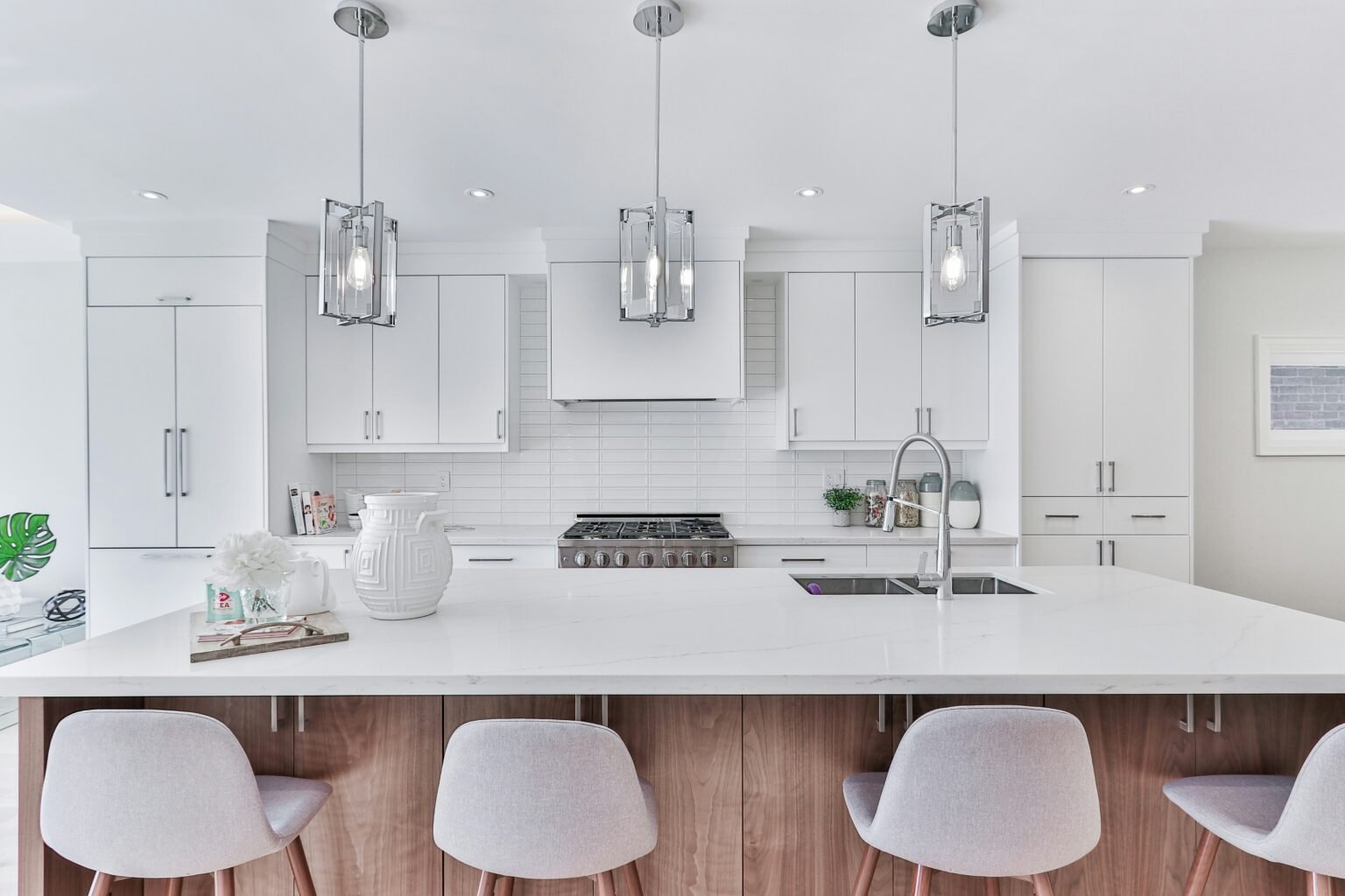 Bright contemporary kitchen featuring a white quartz island with wood paneling, sleek white cabinetry, and stainless steel appliances. The design includes three modern pendant lights, a double-basin sink with a gooseneck faucet, and cushioned bar stools, all set against a clean subway tile backsplash for a fresh, minimalist look. The best kitchen remodeling in Manhattan.