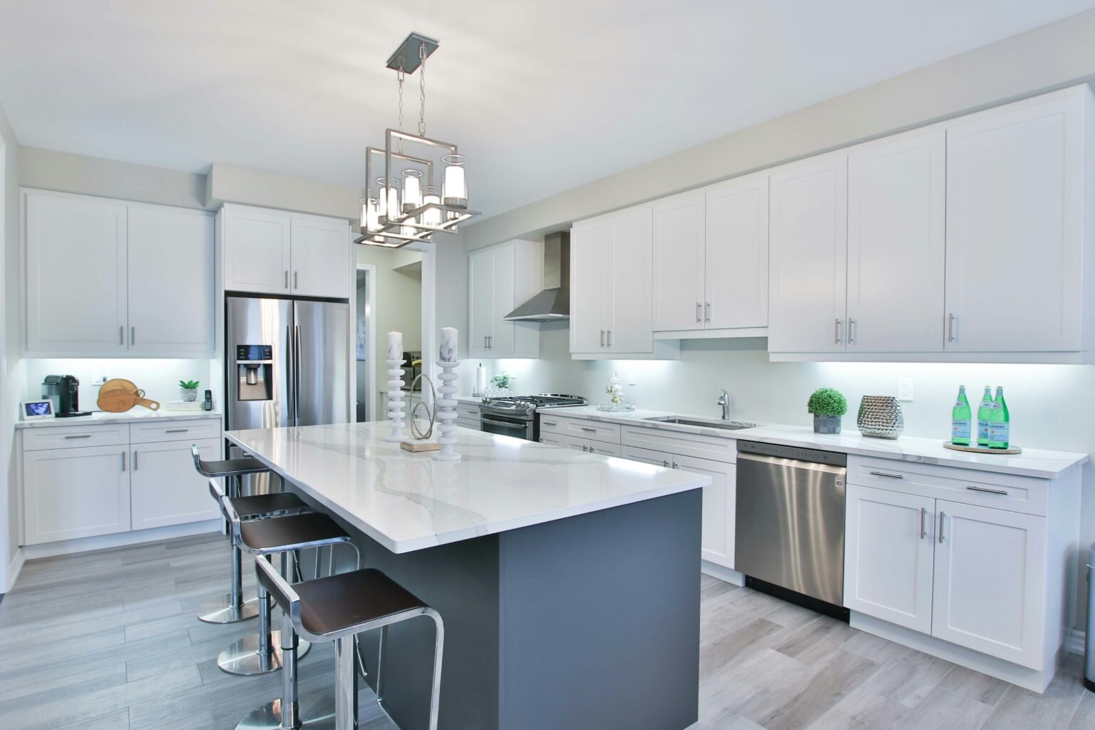 Premier general contractor in Manhattan. Bright modern kitchen with a large white quartz island, sleek gray base, and chrome barstools, featuring white shaker cabinets, stainless steel appliances, under-cabinet lighting, and a contemporary chandelier, blending luxury design with functional elegance.