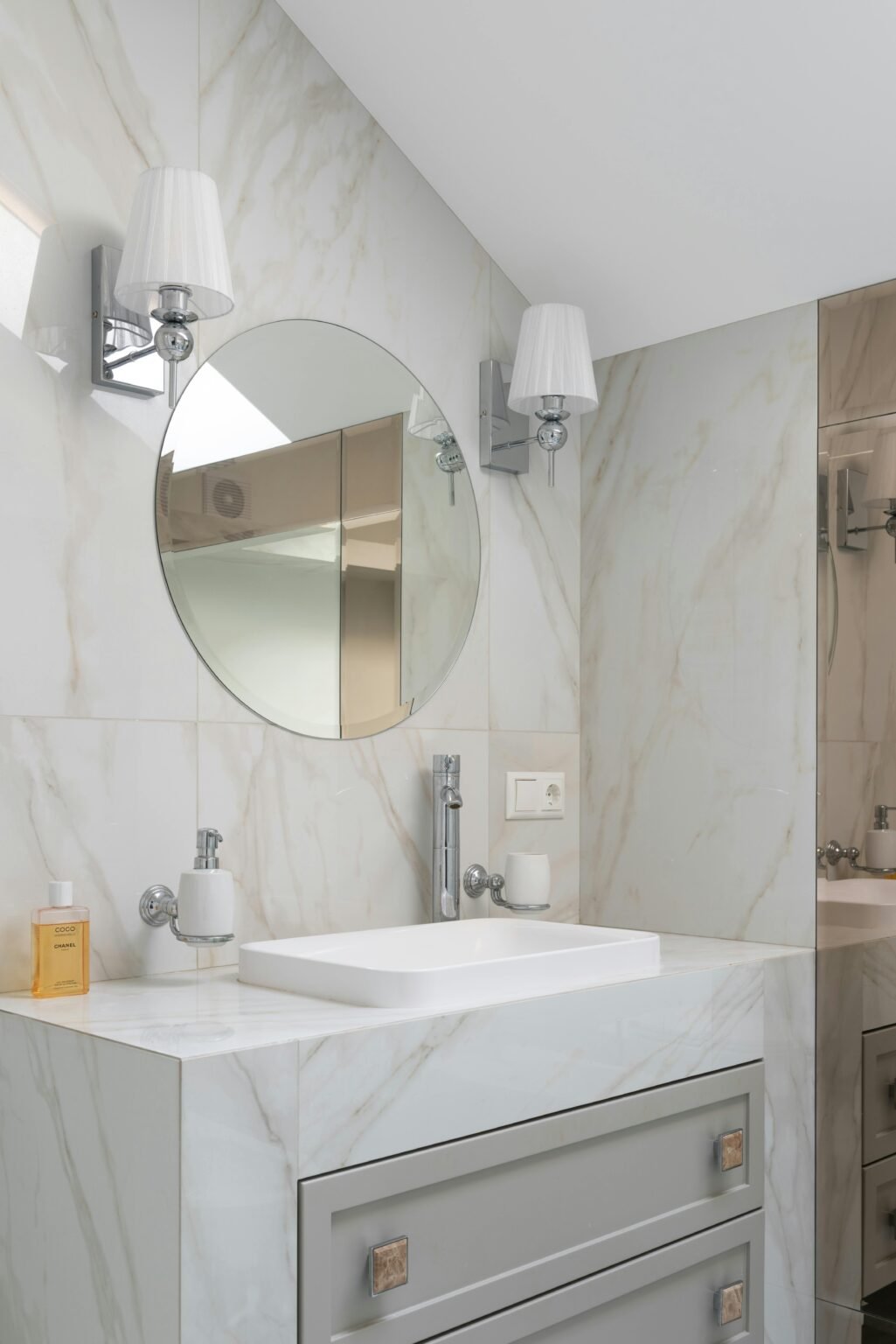 Elegant bathroom vanity with a sleek rectangular sink set in white marble, accented by a round mirror, chrome faucet, wall-mounted soap dispenser, and dual modern sconces with white shades, offering a clean, contemporary design with subtle luxury