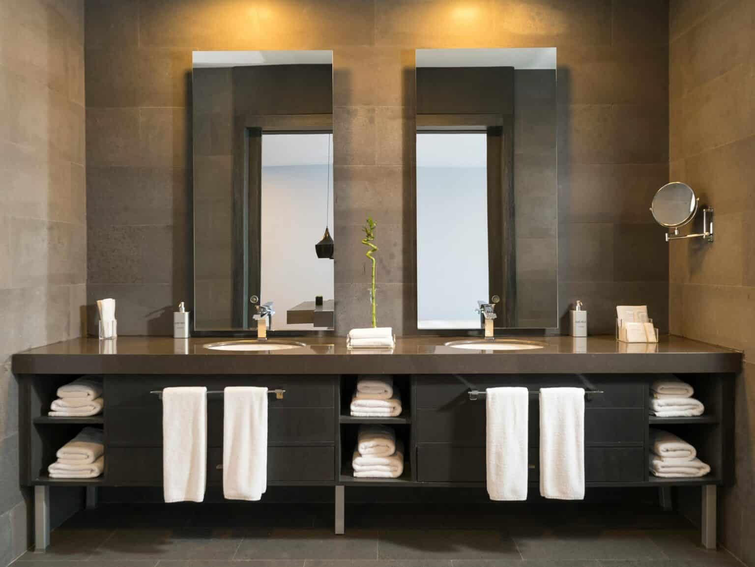 Contemporary bathroom renovation by a general contractor showcasing a double-sink vanity with dark cabinetry, open shelving, and built-in towel storage. Sleek mirrors, warm lighting, and stone tile walls create a luxurious, spa-inspired atmosphere with minimalist appeal.